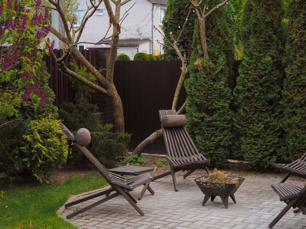 A small backyard with wooden lounge chairs, a fire pit, and a dense evergreen hedge screening the fence line
